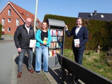 drei Personen mit Buch in der Hand am Bücherschrankk