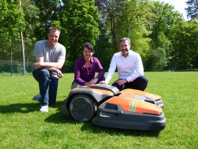 drei Personen posieren mit Mähroboter auf Sportplatzwiese